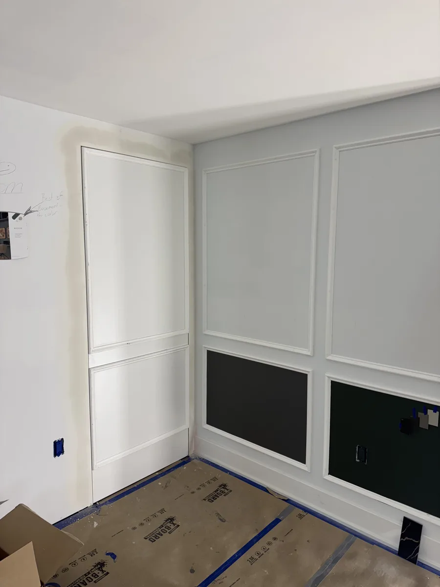 During construction — hidden door panel flanked by picture-frame wall moulding and dark accent cabinet doors, protective floor covering in place